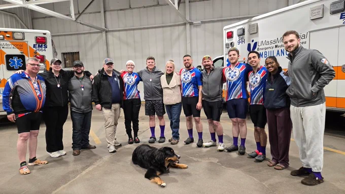 Group of cyclists and emergency personnel standing together inside a garage with two ambulances in the background and a dog lying in front.