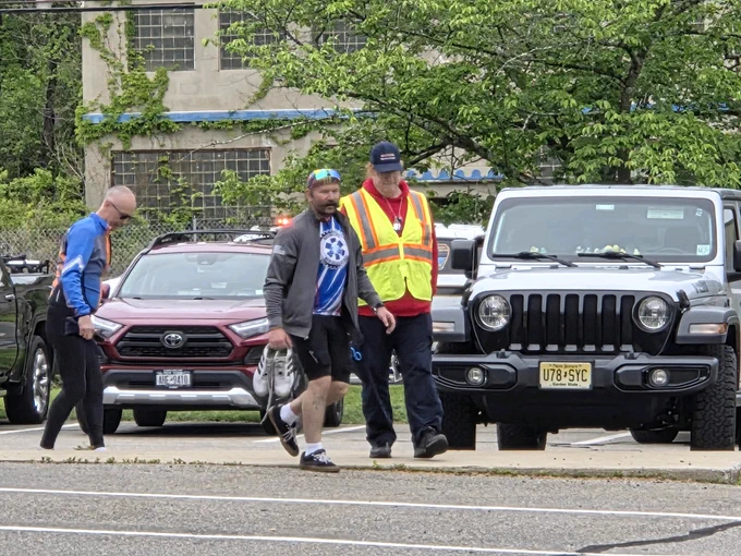 Two men, one wearing a safety vest, walk on a sidewalk near parked cars including a black Jeep and a red Toyota in an urban area with greenery.