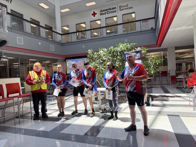 Group of six people standing in a sunlit atrium of the American Red Cross Blood Services building, five wearing matching red, white, and blue cycling jerseys holding papers.