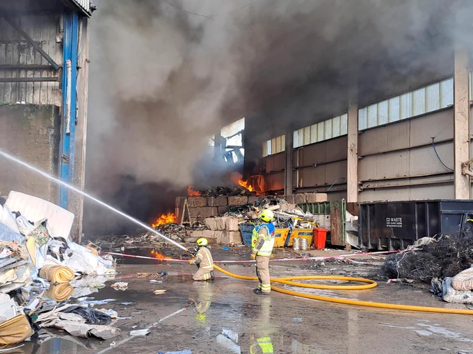 A firefighter on one knee spraying water onto a fire, with another firefighter monitoring behind. In the background, a plume of smoke at a recycling centre.