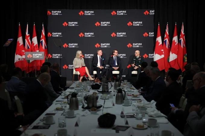 Panel discussion with four speakers seated on stage surrounded by flags and an audience at tables.