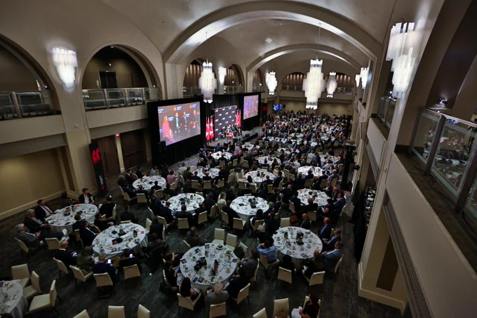 Large conference hall filled with round tables and attendees watching a presentation on two large screens at the front.