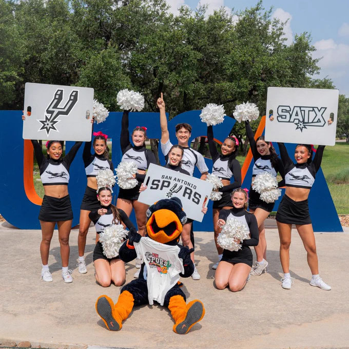 A group of cheerleaders is posing together outside with pom-poms. They are holding signs with the San Antonio Spurs logo and "SATX". A mascot sits in front, and a large "UTSA" sign is visible in the background.
