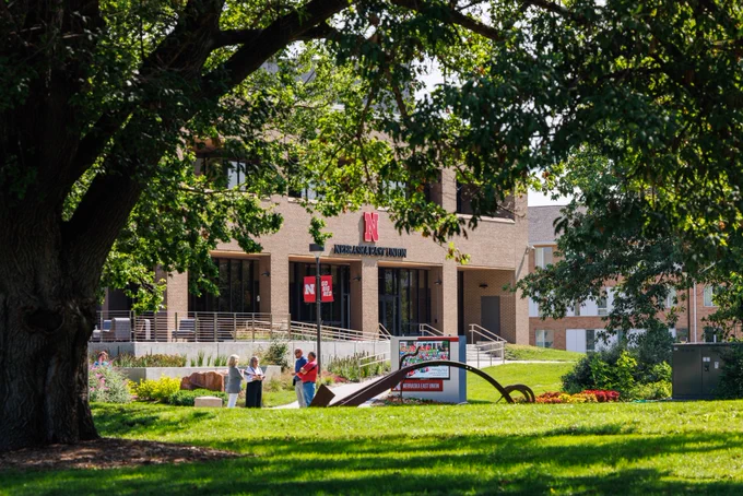 Scenic image of the East Campus Union on a sunny day. The grass is a bright green, and the camera captures foliage covering the brown union building. In the distance is a group of meeting hanging out near a sculpture.