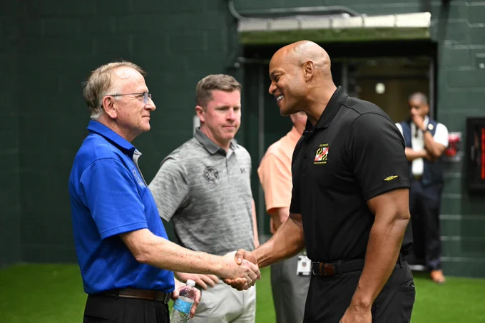 A group of people are indoors, shaking hands and having a conversation at sports training facility with green flooring. One person is wearing a blue polo shirt, holding a water bottle, and another in a black polo shirt with the Maryland state flag logo. 