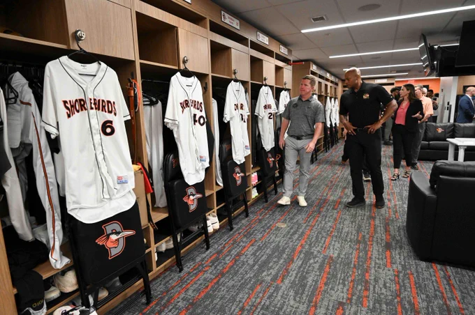 A group of people stand in a baseball locker room, observing the lockers. The lockers contain jerseys with the name "Shorebirds" displayed. 
