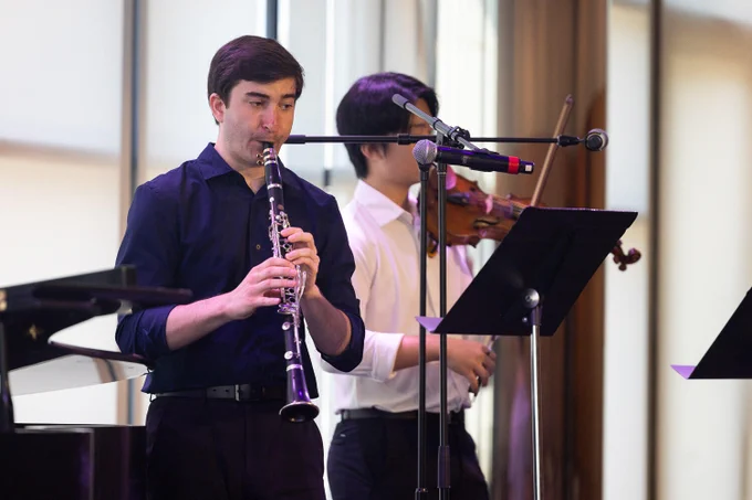 Two individuals are performing music. The person in the foreground is playing a clarinet, and the person in the background is playing a violin. They are standing near music stands and microphones.
