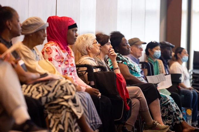 A diverse group of people sit attentively in a row, possibly at a public event or meeting. Some wear face masks, and most appear engaged with the speaker or presentation.