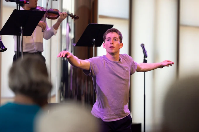 A person in a casual outfit performs a dance in front of a microphone stand, with a violinist playing nearby. The background includes an audience and soft lighting.