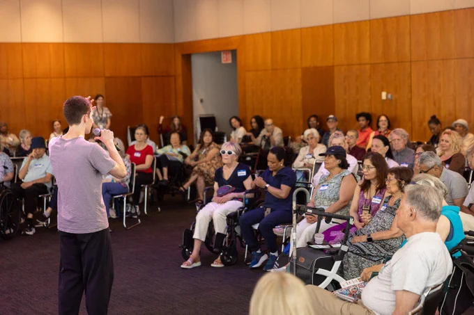 A person is speaking to a seated audience in a community event space with wooden walls. The attendees are diverse in age and appear attentive. Some are seated in wheelchairs. The atmosphere is casual and engaged.