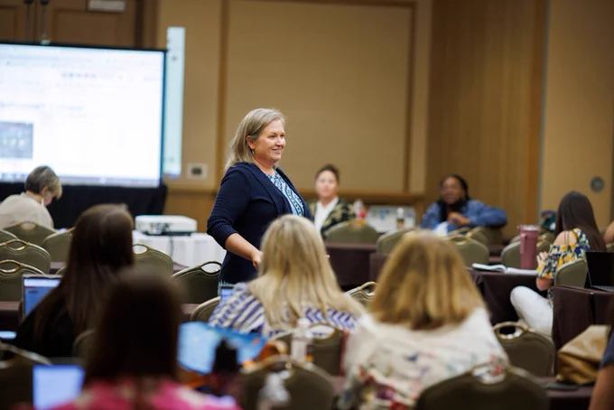 Photo of a person standing in a room full of people who are sitting and watching. The person is seems to be teaching, with a screen behind them showing information. 