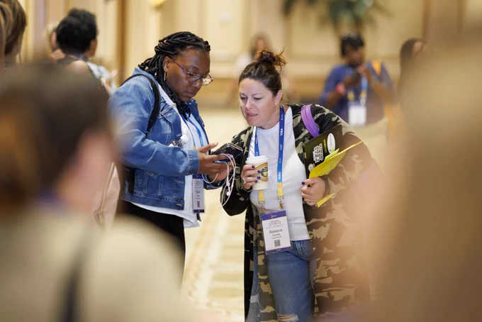 Photo of two people looking at something on one of their phones. They are standing in a hallway with other people passing by. 