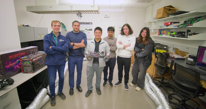 Group of six people standing in a lab with one person holding a robotic vehicle model in the center.