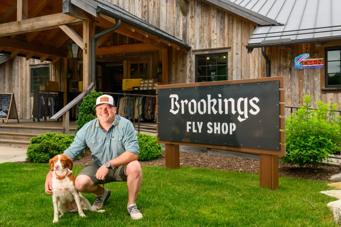 A person kneels beside a dog in front of Brookings Fly Shop. The building exterior is wooden, with a visible "Open" sign in the window.