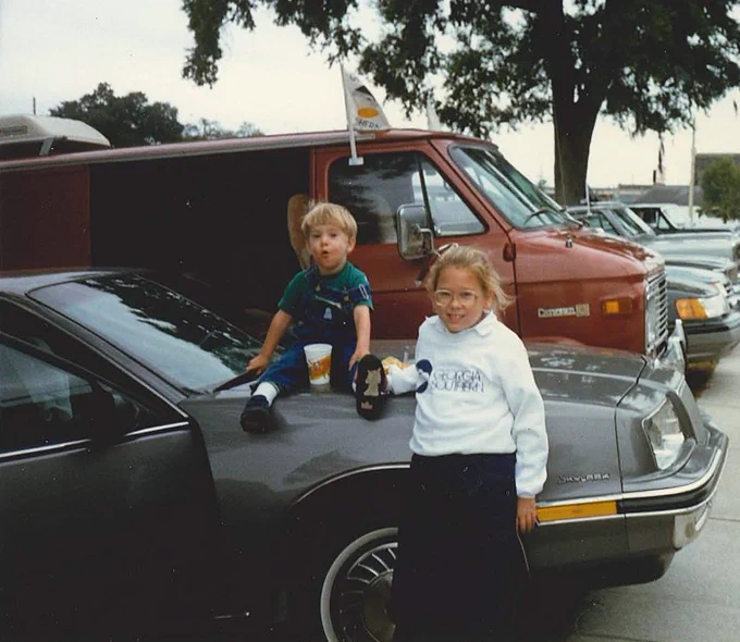 Jennifer and her brother at a Georgia Southern away game in Jacksonville, FL, in 1987. As children, her brother sits on the hood of a car as she stands beside the car.