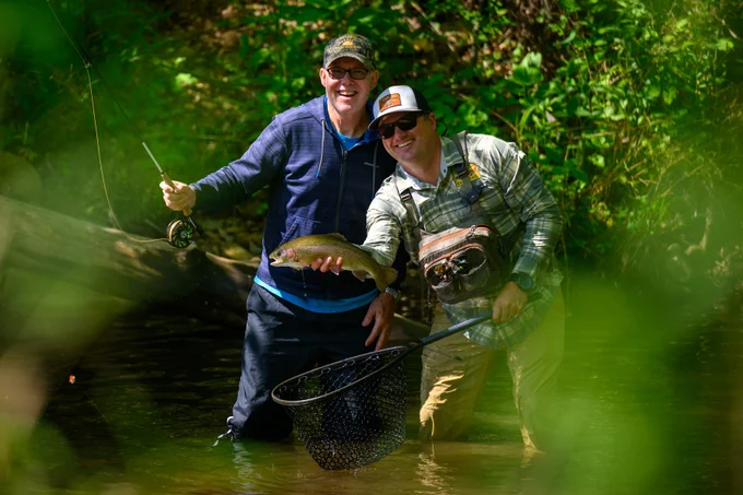 Two people stand in a shallow stream surrounded by greenery, smiling and holding a fishing net containing a fish. They appear to be enjoying a successful fishing trip.