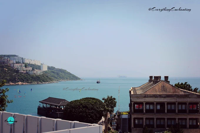 An elevated, scenic landscape view of Repulse Bay in Hong Kong, captured in the image. Calm turquoise ocean waters stretch out under a clear, pale blue sky, with small boats and sailboats visible in the distance. To the left, a lush green hillside is dotted with high-rise buildings, while a traditional covered wooden pier extends into the bay. In the right foreground, a multi-story building with a tiled roof stands surrounded by dense green trees. Taken by UK travel blogger Anamika Chattopadhyaya for her blog 'Everything Enchanting'.