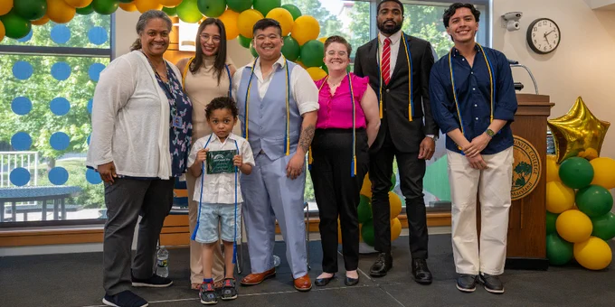 Group of six adults and one child posing indoors with green and yellow balloon arch in background.