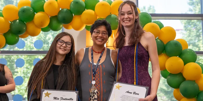 Two students holding certificates stand beside a faculty member in front of a green and yellow balloon arch indoors.