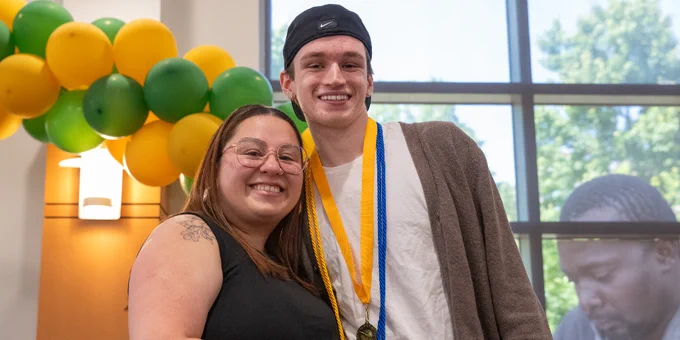 Two people posing indoors near a green and yellow balloon arch, one wearing a medal and honor cords around their neck.