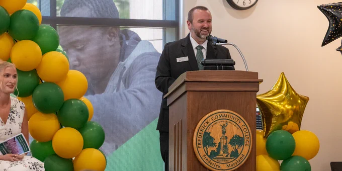 AVP for Student Engagement and Wellness, Zach Roberson, in formal attire delivering a speech at a podium decorated with Frederick Community College emblem and surrounded by green and yellow balloons.