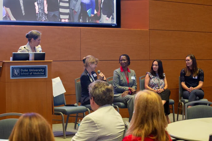 Panel discussion with four seated participants on stage and one moderator at a podium; audience members sit at tables in the foreground watching the discussion.