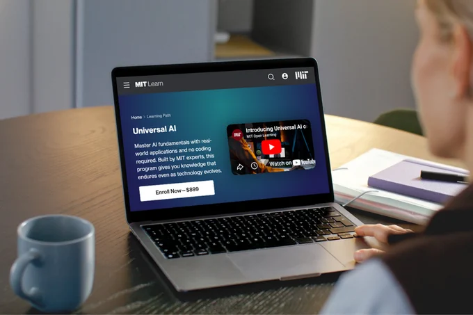 Laptop displaying a website titled "MIT Learn" with a course on "Universal AI." The screen shows a video thumbnail and a description of the course, offering enrollment for $999. A table with notebooks and a coffee mug are nearby.