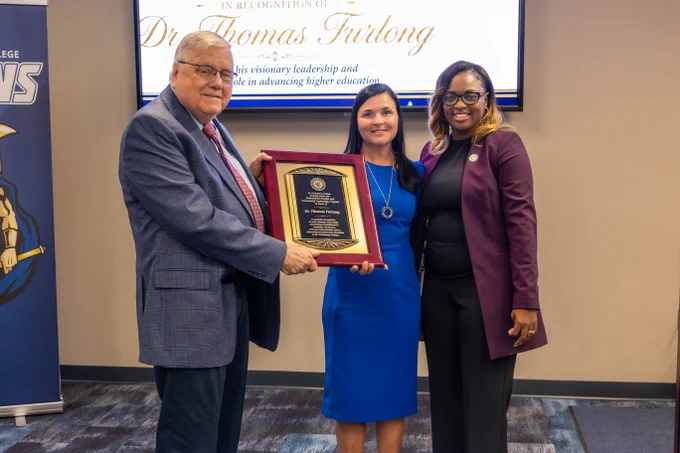 A group of three individuals stands together in a formal setting. One person is holding a plaque. A backdrop displays the text "In recognition of Dr. Thomas Furlong" and references contributions to SPC and higher education.