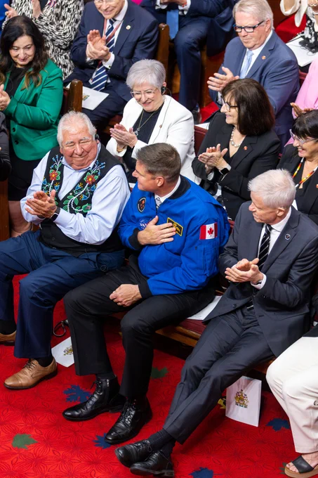 Des membres du public assis et qui applaudissent lors d’un événement formel, dont un homme portant une veste bleue avec un écusson du drapeau canadien.