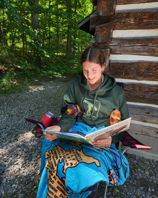 Person in a green Ontario Parks hoodie, sitting on a camp chair, with two stuffed beavers reading a book. They have an Ontario Parks blanket on their lap.