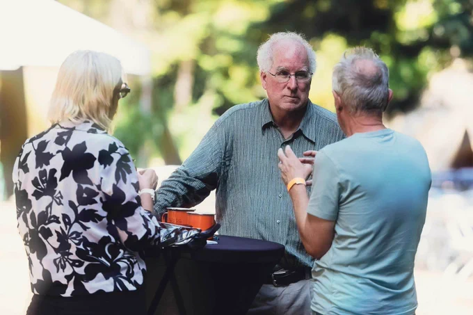 Brewster Kahle, wearing a blue-gray button-up shirt and glasses, listens attentively in conversation with two other people at an outdoor standing table at DWeb Camp. One person faces away wearing a black-and-white floral blouse; another faces away in a light teal t-shirt with an orange wristband.