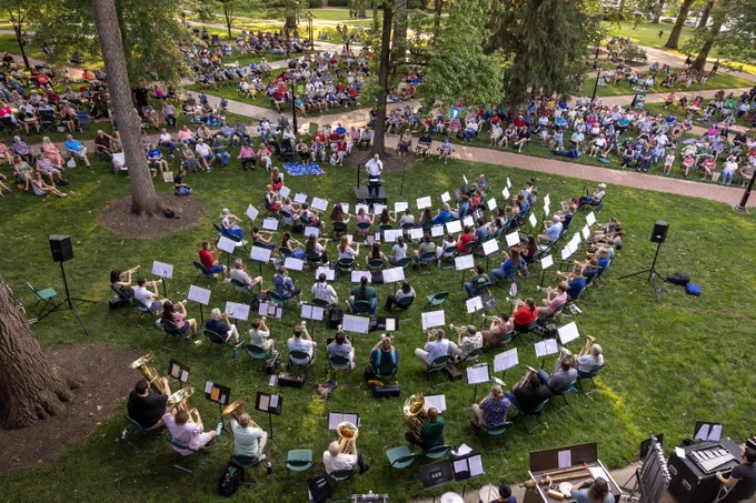 An outdoor concert with a large orchestra performing in a park surrounded by a seated audience. Trees and pathways can be seen in the background.
