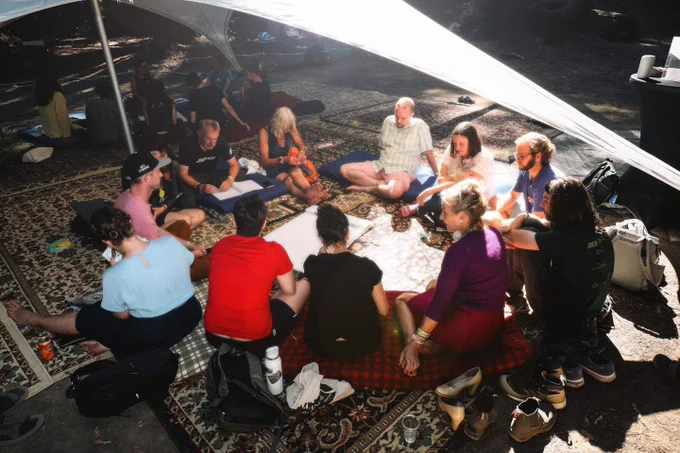 A group of roughly a dozen people sit in a circle on colorful rugs and cushions under a large white canopy tent at DWeb Camp, engaged in discussion. Papers and notebooks are spread in the center. Sunlight filters through the tent fabric.