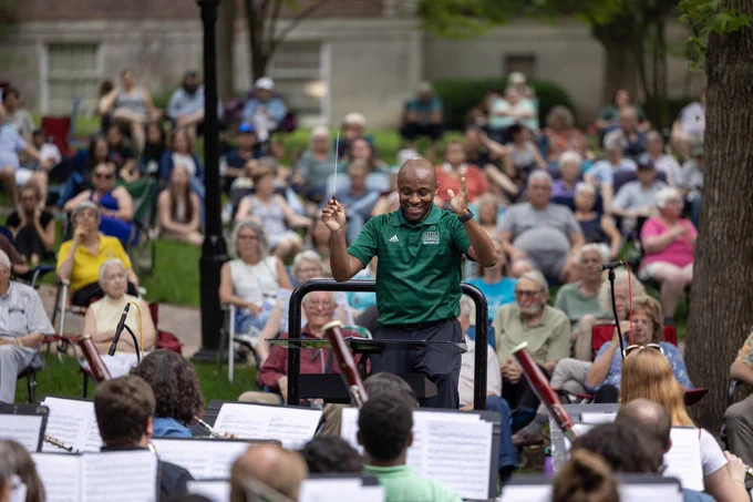Outdoor concert with a conductor leading an orchestra in front of a large seated audience in a park setting. Trees and a building are visible in the background.