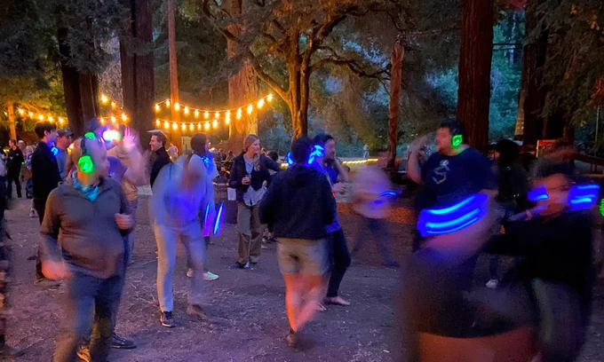 A crowd of people dance outdoors at night among towering redwood trees at DWeb Camp's silent disco. Many wear glowing LED headphones in blue and green. String lights hang between the trees, and motion blur conveys the energy of the dancing crowd.
