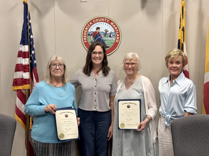 Four individuals stand side by side in an indoor government meeting room, positioned in front of a wall displaying the Frederick County, Maryland seal at the center. On each side of the seal, two flags are visible: the United States flag on the left and the Maryland state flag on the right. The individuals are dressed in professional clothing, and two of them hold framed Certificates of Appreciation with official seals and printed text. The group stands between two gray upholstered chairs that are partially visible at the edges of the image. 