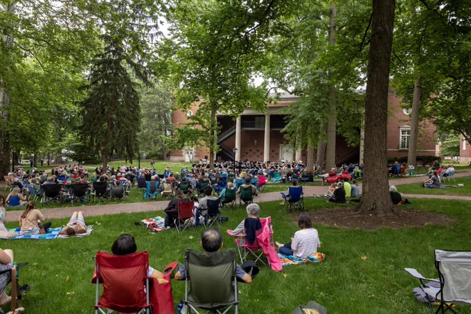 A large group of people is seated outdoors, some on chairs and others on the grass, attending a concert at an amphitheater surrounded by lush trees. The atmosphere is relaxed, with a stage in the distance.