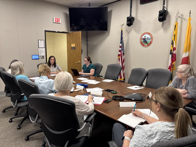 A group of people sits around a large rectangular conference table in a government meeting room. The table is covered with papers, notebooks, folders, and a central conference speakerphone. Several individuals are writing in notebooks or reviewing documents, while one person uses a laptop. The walls of the room are plain and light-colored, with the Frederick County, Maryland seal mounted on the wall behind the table. To the right of the seal stand three flags on poles: the U.S. flag, the Maryland state flag, and another flag with red and yellow sections. A closed wooden door is visible on the left side of the room beneath an illuminated EXIT sign. A wall-mounted television screen hangs above the conference area, and multiple office chairs with dark upholstery surround the table. 