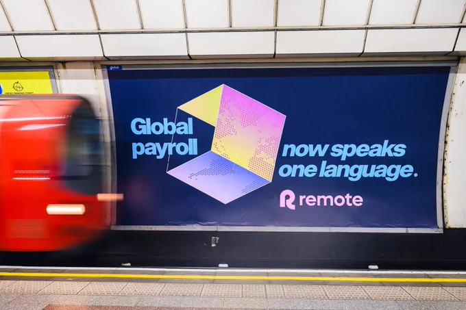 Remote global payroll platform billboard on London Underground platform featuring blue background with 3D geometric design and headline "Global payroll now speaks one language" promoting payroll infrastructure for global teams.