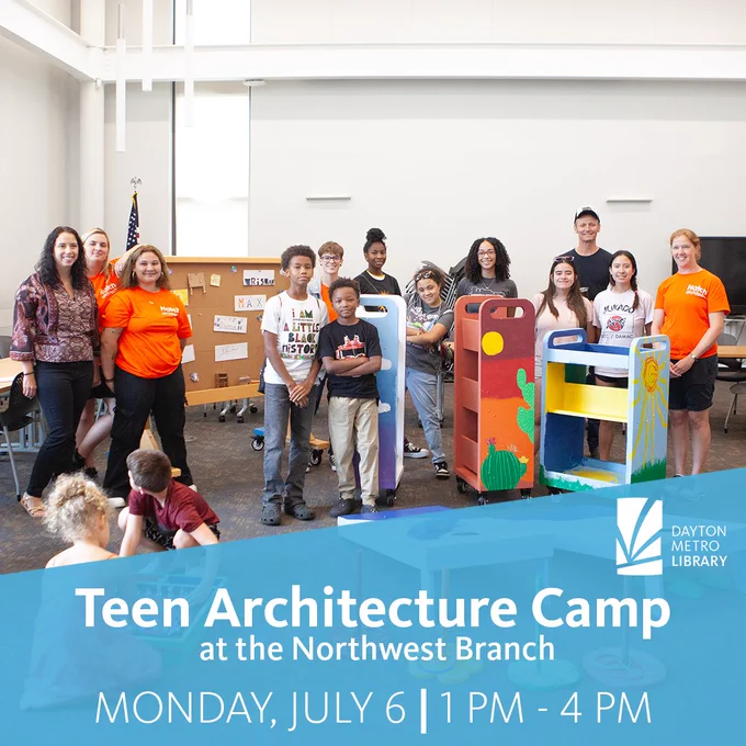 A group of teens and adult instructors stand inside a bright library space behind several colorful, hand painted rolling carts. The carts feature designs like a desert scene and a sunrise. A corkboard with sketches and notes is visible in the background.