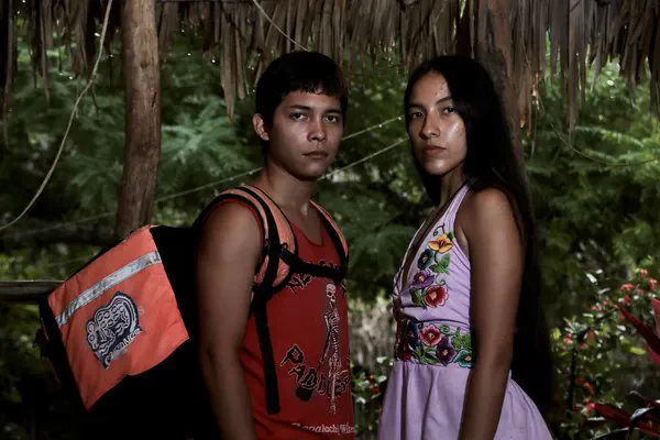 A still from the film Wetiko. A Mayan teenage boy carrying a cube-shaped backpack stands opposite a young Mayan woman in the jungle. Both are looking very directly to camera.