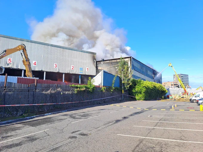 Photo taken from a car park showing a plume of smoke over two industrial units at a recycling centre. On the right hand side in the background, a turntable ladder sprays water onto the building.