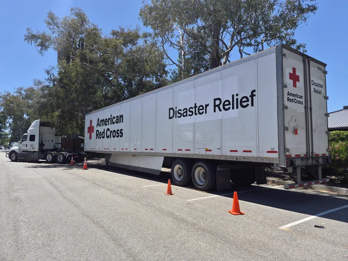 A large American Red Cross Disaster Relief truck is parked on the side of the road, adjacent to some trees. Several orange traffic cones are placed near the truck.