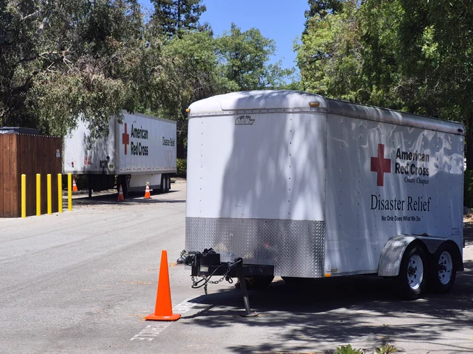 Two American Red Cross disaster relief trailers are parked on a paved path surrounded by trees. Bright orange traffic cones are positioned near the trailers.
