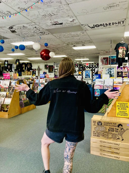 A person with tattooed legs stands in a vibrant record store, gesturing with open arms. The ceiling is covered with graffiti and signatures, adding an artistic touch.