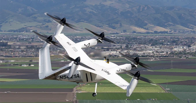 A white Joby Aviation aircraft with multiple rotors is flying over a rural landscape with fields and hills. It is a vertical takeoff and landing vehicle, designed for advanced air mobility.