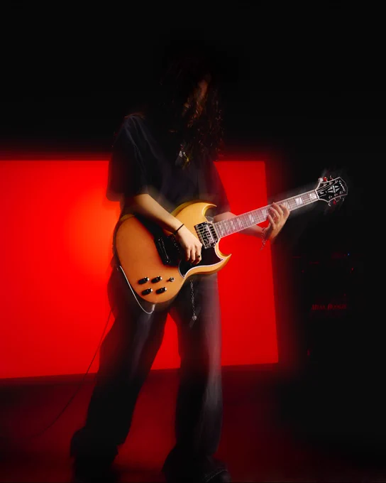 A person plays an Epiphone Futura SG Custom lectric guitar in front of a bright red background. The scene is dimly lit, with the guitar and red light providing contrast. A "Mesa Boogie" amplifier is partially visible in the background.