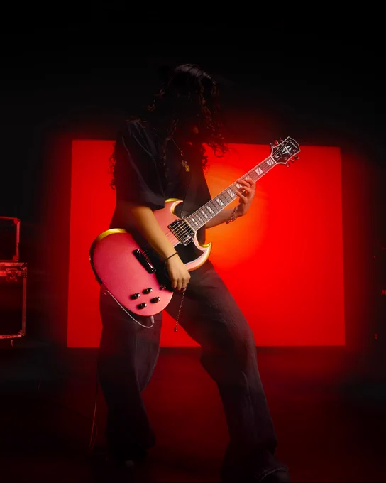 A person playing an Epiphone Futura SG Custom electric guitar in front of a vivid red backdrop. The scene is dimly lit, creating a dramatic effect with the glowing background highlighting the subject. The guitar is pink and the person’s hair is long, cascading over their face. A blurred amplifier is visible on the side.