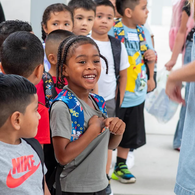 A young student wearing a colorful backpack smiles on the first day of school surrounded by classmates.