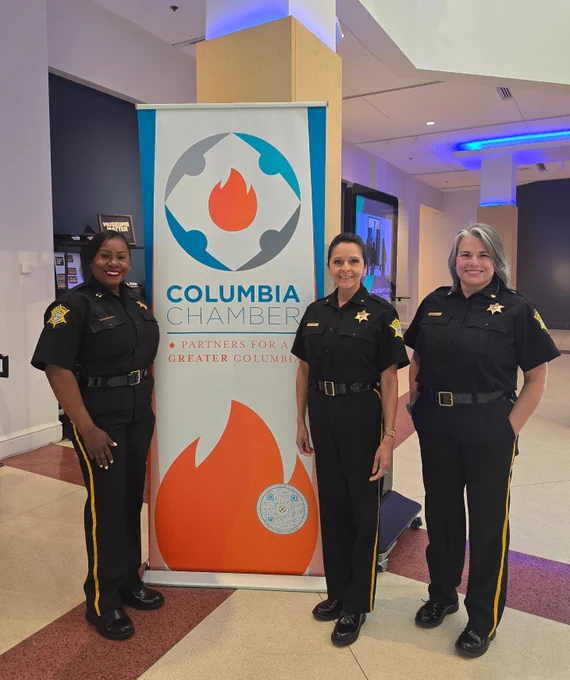 Three people in sheriff uniforms stand smiling in front of a Columbia Chamber banner inside a building.
